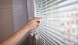 A person's hand delicately wiping dust off white blinds with a rag, representing the meticulous work of professional cleaning services that can contribute to a clean and peaceful living space.