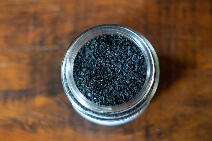 A glass jar filled with small, porous activated charcoal pellets on a wooden surface, used for its natural odor-absorbing properties to freshen air from smoke odors.