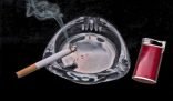 A lit cigarette resting on a clear glass ashtray beside a red lighter, emitting smoke that symbolizes the challenge of eliminating smoke odor in the home.