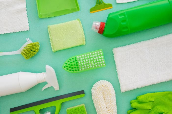 An assortment of green cleaning tools and products, including sponges, brushes, gloves, and a spray bottle, arranged on a bright turquoise background.
