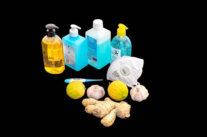 A selection of disinfectants and natural remedies including hand sanitizer, antiseptic liquids, a mask, and fresh ginger, garlic, and lemons, representing a comprehensive approach to home disinfection.