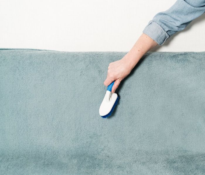 Close-up of a hand using a fabric brush on a blue sofa, demonstrating a step in the deep cleaning process for maintaining the sofa's cleanliness and hygiene.