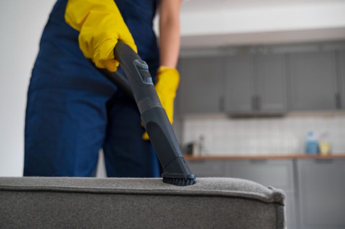 A professional cleaner in a blue uniform and yellow gloves vacuuming a grey fabric sofa, highlighting the process of assessing upholstery fabric before deep cleaning.