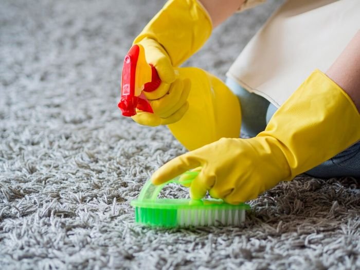 A person in yellow gloves testing a cleaning solution on a grey carpet, ensuring the safety and effectiveness before broader application.