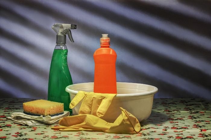 Essential cleaning tools, including spray bottles, detergents, a basin, sponge, and gloves, arranged on a floral-patterned surface, ready for an efficient top to bottom, left to right cleaning strategy.