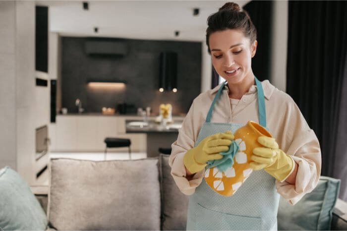 A smiling woman with a neat bun and apron happily cleans a child's toy with a yellow sponge, wearing rubber gloves. This showcases the involvement of family members in home cleaning tasks, fostering teamwork and responsibility.
