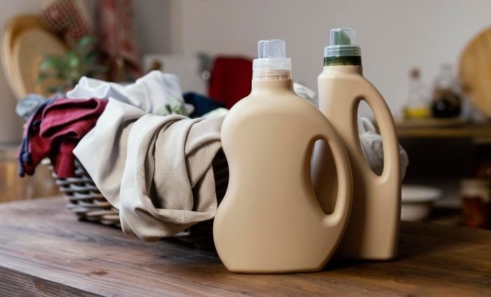 Two eco-friendly laundry detergent bottles on a wooden surface with a basket of clothes in the background, representing DIY solutions for removing tough grass stains.