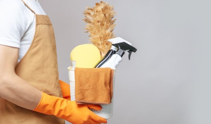 Person clad in a beige apron and orange gloves holding a variety of cleaning tools and products, including a feather duster, spray bottle, and sponges, signifying readiness for home cleaning tasks.