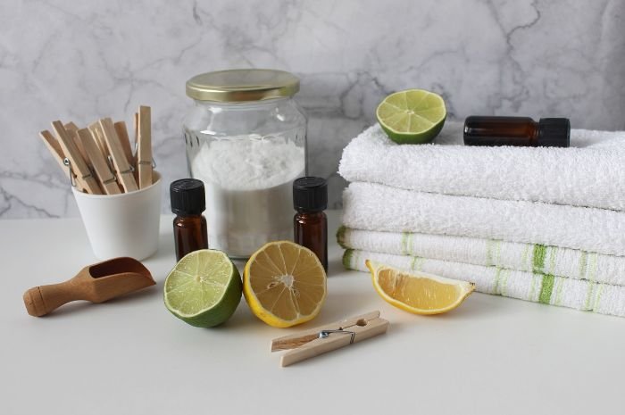 Eco-friendly laundry essentials on a marble countertop, including a glass jar of baking soda, fresh lemons and lime, wooden clothespins, essential oil bottles, and white towels, ready for creating all-purpose, natural stain removers.