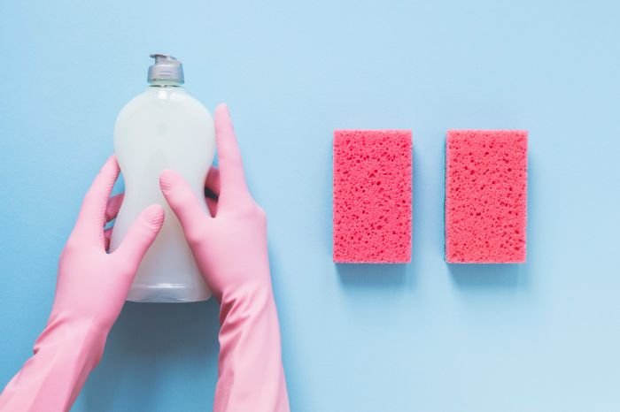 Hands in pink rubber gloves holding a clear bottle of liquid next to two pink sponges on a light blue background, symbolizing the initial steps in pre-treating oil stains on fabrics.