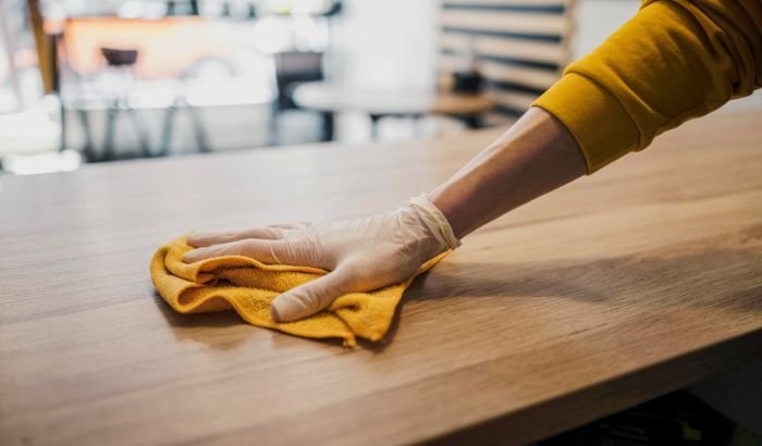 A person's hand in a protective glove wiping a wooden table with a yellow cloth, highlighting a tip for removing stains from wooden furniture.