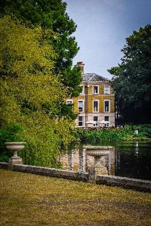 A tranquil view of a classic house overlooking a pond surrounded by lush greenery, demonstrating the efficiency and value of professional landscaping and design in creating harmonious and beautiful outdoor spaces.