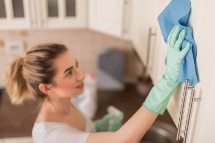 A woman in casual attire efficiently wipes down a stainless steel refrigerator with a blue microfiber cloth while wearing protective green gloves, exemplifying a quick and effective daily cleaning routine.