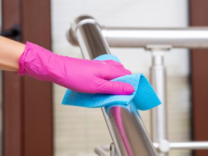 Hand in pink glove wiping a stainless steel railing with a blue cloth, demonstrating the use of a homemade cleaner with olive oil and vinegar to restore the shine on stainless steel surfaces.