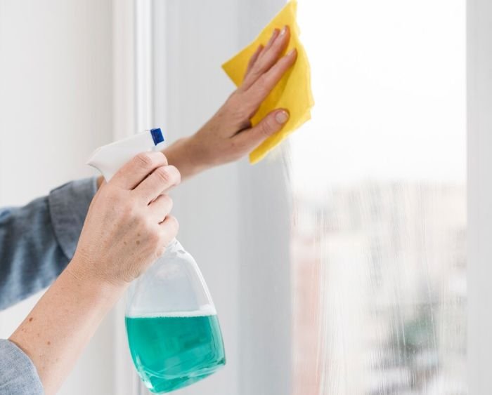 Close-up of hands using a homemade window cleaner solution, spraying it onto a window and wiping with a yellow cloth, demonstrating an effective, natural cleaning method.