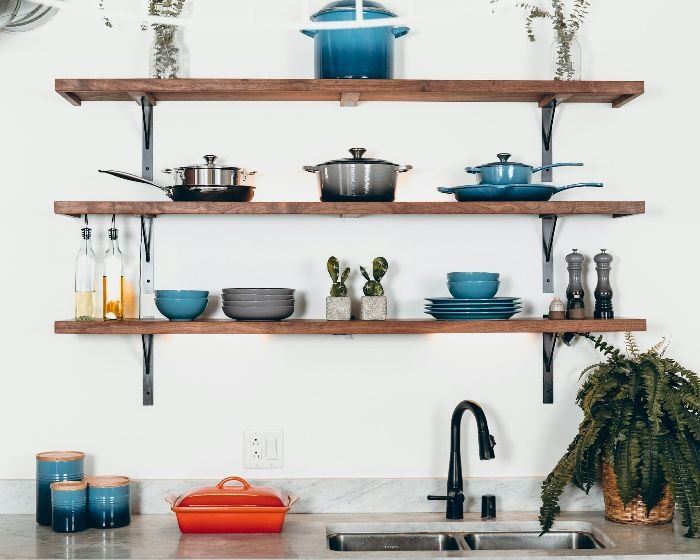 A tidy kitchen area showcasing well-organized open wooden shelves filled with various pots, pans, and dishes in shades of blue and gray, with cooking oils and a potted plant, emphasizing an efficient and aesthetically pleasing storage solution.