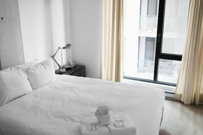 A well-organized bedroom featuring a neatly made bed with a decorative towel fold on white linens, a modern bedside lamp, and a view of a city through a floor-to-ceiling window, embodying a clean and serene morning environment.