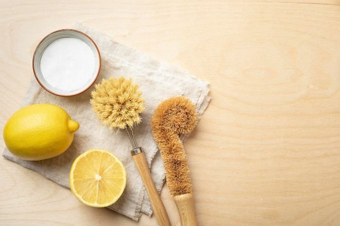 Natural cleaning supplies for bird cage maintenance, including a lemon, baking soda, and wooden brushes, emphasizing safe, non-toxic products.