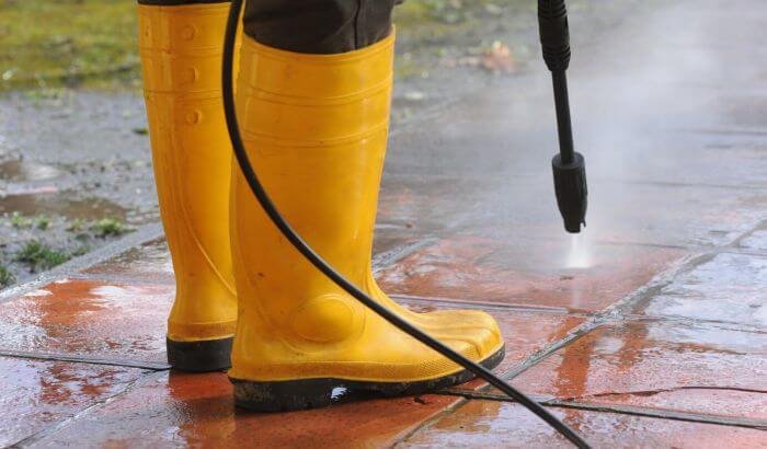 Person wearing bright yellow rubber boots using a pressure washer to clean a brick patio, demonstrating effective outdoor cleaning techniques for decks and patios.