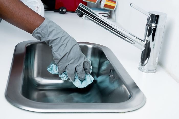 A professional cleaner wearing gloves and cleaning a stainless steel sink, illustrating the benefits of hiring professional cleaning services to save time and maintain a fresh home.
