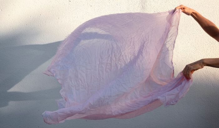 Hands gently holding and waving a delicate pink silk fabric in the sunlight against a textured white wall, symbolizing care for fine textiles.