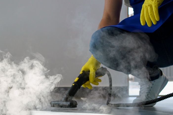 A person in blue overalls and yellow gloves uses a steam cleaner on a floor, demonstrating the effective removal of dirt through DIY deep cleaning techniques, emphasizing the efficiency of steam in penetrating and cleaning floor surfaces.