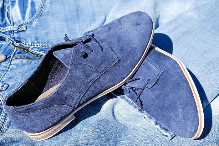 A pair of blue suede shoes resting on a denim background, illustrating the unique texture and characteristics of natural suede susceptible to stains, ideal for discussing suede care in a blog post.