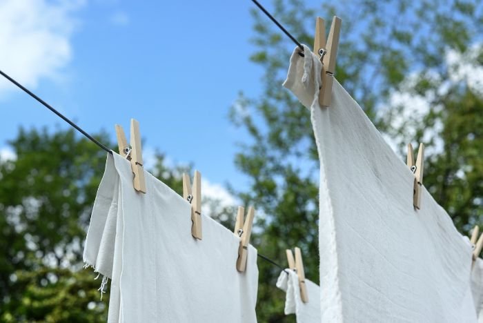 White clothing hung to dry on a clothesline outdoors, secured with wooden pegs against a backdrop of blue sky and green trees, highlighting effective drying practices for maintaining fabric quality.