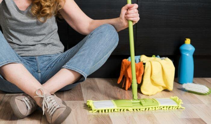 A person sitting on the floor, taking a break from cleaning, holding a mop handle, with cleaning gloves, cloths, and detergent bottles around her, emphasizing the tools and efforts involved in weekly deep cleaning for maintaining a clean home.