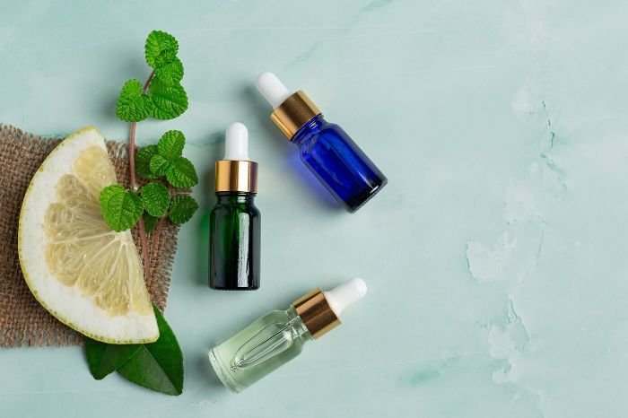 Three essential oil dropper bottles, one green, one blue, and one clear, are placed on a light green surface alongside a lemon slice and a sprig of mint, representing essential oils that are safe and those to avoid around children.