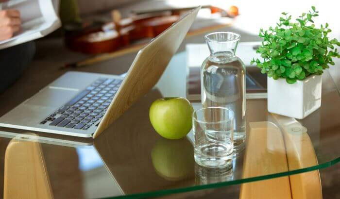 A clean glass table with a laptop, an apple, a water carafe, a glass, and a potted plant, illustrating the results of deep cleaning windows and glass surfaces.