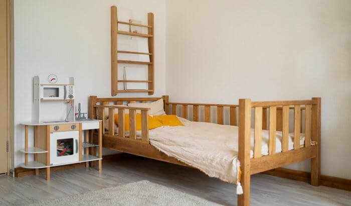 Wooden toddler bed with beige bedding in a minimalistic children's room, accompanied by a toy kitchen set.