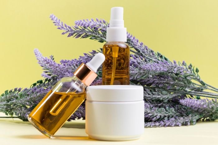 Bottles of essential oils and a white container are arranged in front of lavender sprigs, showcasing ingredients for making a natural disinfectant spray.