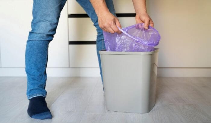 A person placing a purple trash bag into a gray trash bin, illustrating the process of cleaning and removing stains from plastic items.