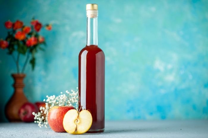 A bottle of vinegar with apples and flowers in the background, illustrating the use of vinegar as an effective pre-treatment solution for removing stains from plastic items.