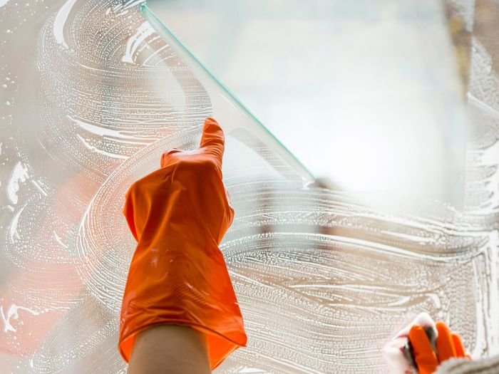 A close-up of a person wearing orange gloves using a squeegee to clean a soapy window, demonstrating the technique for achieving streak-free windows.