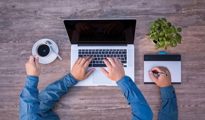 Person multitasking with a laptop and a cup of coffee on a wooden table, illustrating the concept of multitasking while cleaning.