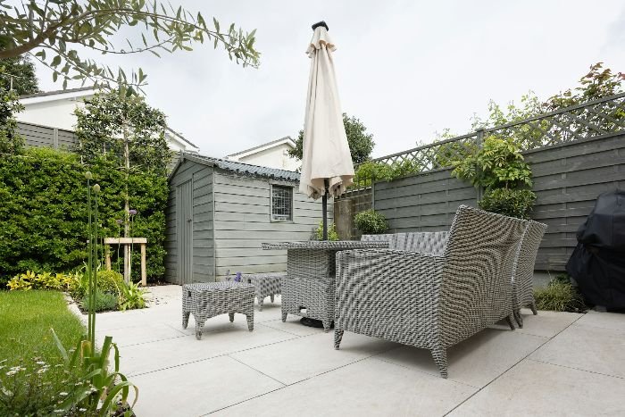 Stylish outdoor patio with wicker furniture, including chairs and a dining set, complemented by greenery and a garden shed, creating a comfortable and inviting space for relaxation.