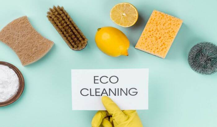 Assortment of natural cleaning products and a hand holding a 'ECO CLEANING' sign on a teal background to illustrate eco-friendly cleaning methods