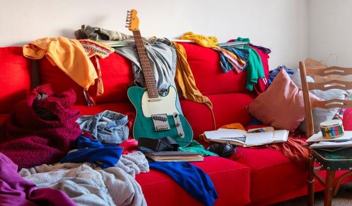 A cluttered living room with clothes, a guitar, and various items scattered on a red couch, illustrating the need for seasonal decluttering.