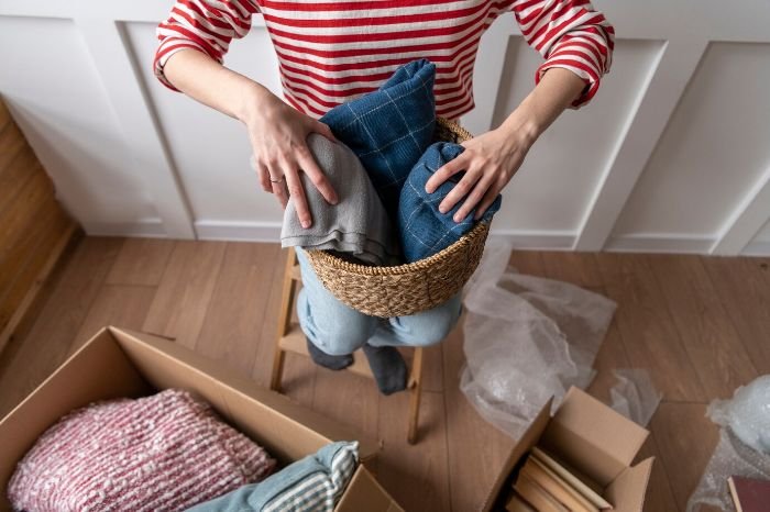 Person sorting seasonal decor by placing folded blankets into a woven basket, with cardboard boxes nearby, illustrating the process of organizing autumn decorations.