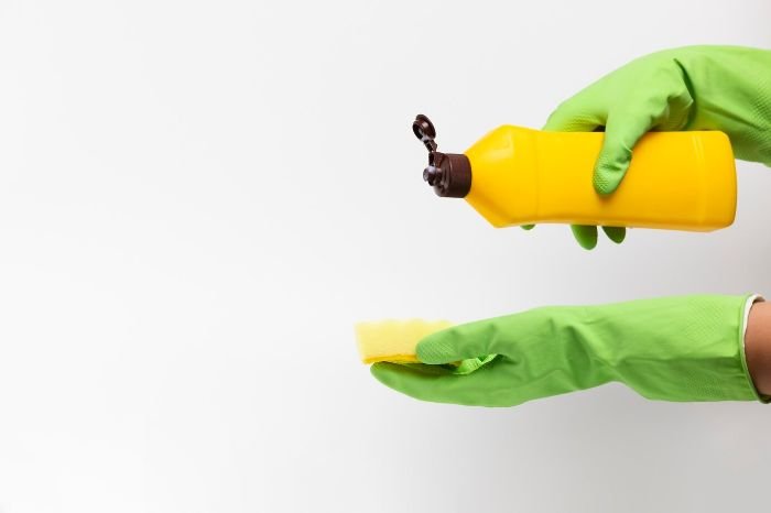 Gloved hands holding a yellow bottle of non-toxic cleaner and a sponge, highlighting the importance of using safe, eco-friendly cleaning products that are pet-friendly and effective.