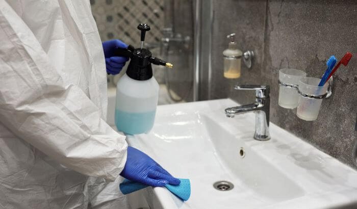 Person disinfecting a bathroom sink with cleaning spray and cloth to prevent illness and maintain hygiene.