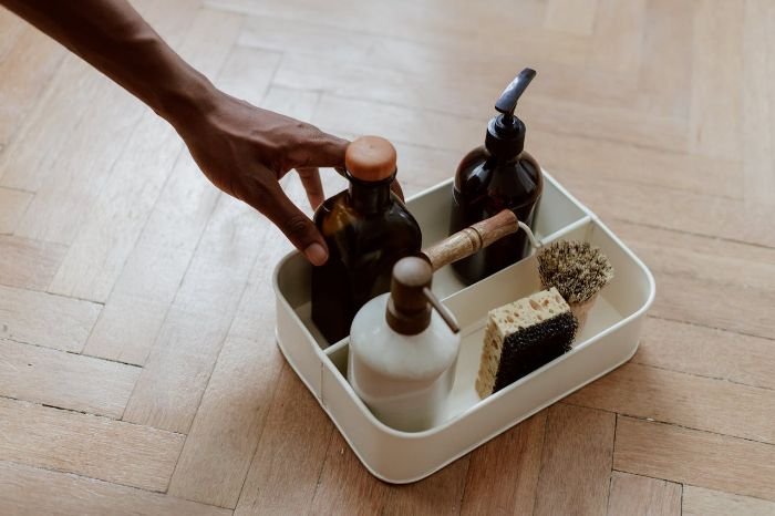 Hand reaching for a cleaning caddy with eco-friendly bottles and brushes on a wooden floor