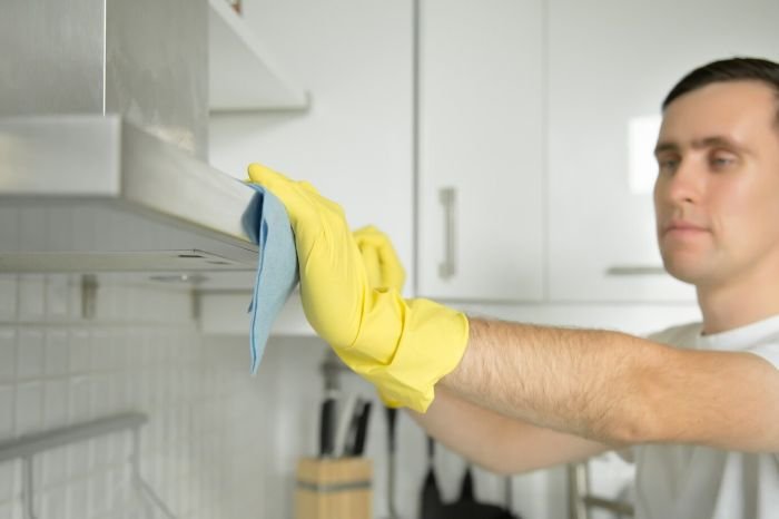 Man wearing yellow gloves cleaning a kitchen appliance with a cloth, demonstrating proper maintenance and cleaning schedules for common kitchen appliances.