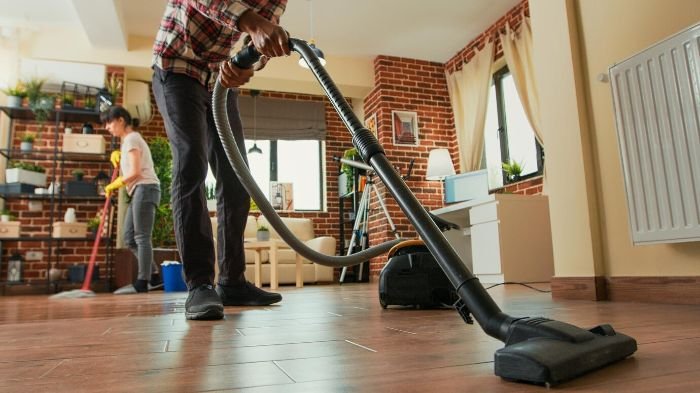 Person vacuuming a hardwood floor with another person mopping in the background, illustrating the 'begin dry, then go wet' cleaning method.