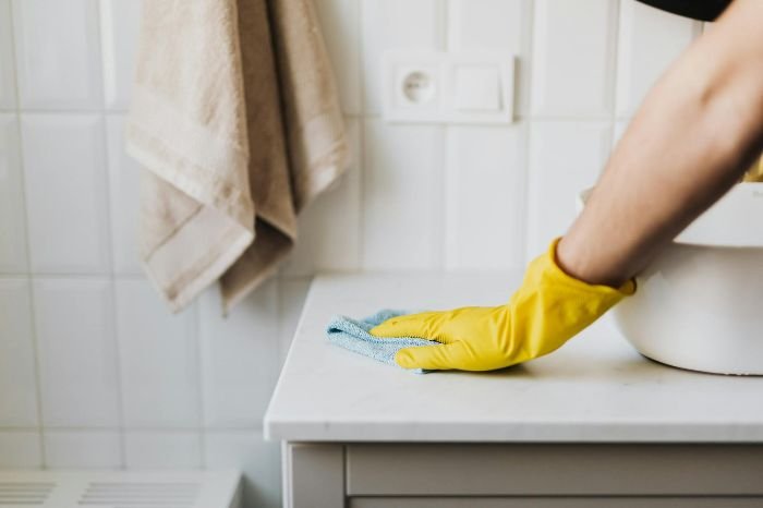 A person wearing yellow rubber gloves cleaning a bathroom countertop with a microfiber cloth, promoting hygiene and routine cleaning benefits.